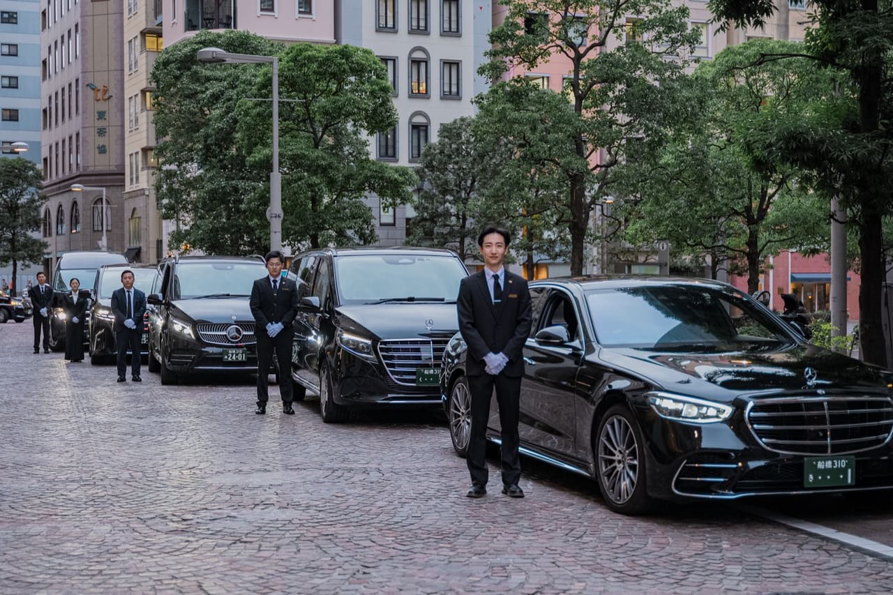 RydAgent chauffeurs in suits standing beside a fleet of black Mercedes and Alphard vehicles on a Tokyo street